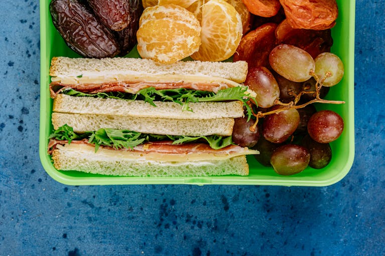 Colorful lunchbox containing ham sandwich, fresh grapes, peeled oranges, and dates on a blue background.