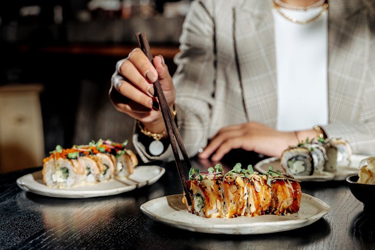 Woman using chopsticks to eat sushi roll with garnish on ceramic plate.